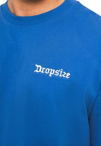 Modrá bavlnená tričko s okrúhlym výstrihom, na prednej strane s bielym textom "Dropsize" v štýlovom písmene, hladká textúra a pohodlný strih.