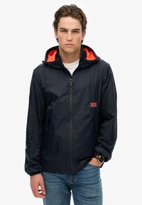 Chaqueta impermeable color marino con detalles en naranja, cierre de cremallera, capucha y bolsillos laterales. Textura suave, diseño ligero apto para uso al aire libre.