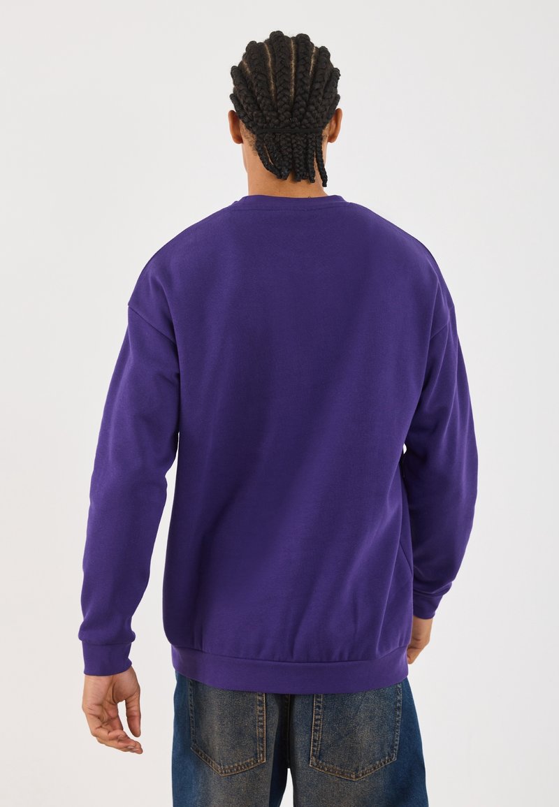 Personne aux cheveux tressés portant un sweat-shirt violet à manches longues et un jean bleu, vue de dos sur un fond uni.