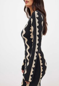 Robe noire ajustée avec un motif beige ondulé, manches longues et texture lisse. La vue de profil met en valeur la silhouette de la robe.