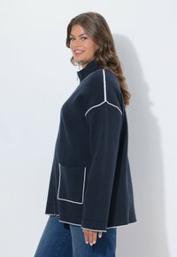 Giacca navy in pile con zip e cuciture bianche, dotata di collo alto, tasca laterale e maniche a vestibilità rilassata che si allargano leggermente.