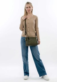 Borsa a tracolla verde oliva con cerniere, texture liscia e tracolla regolabile, abbinata a un cardigan beige a coste e jeans blu.