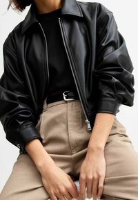 Blouson aviateur en simili cuir noir avec col à revers et fermeture éclair. Caractérisé par des manches amples et une texture lisse. Associé à un pantalon beige taille haute.