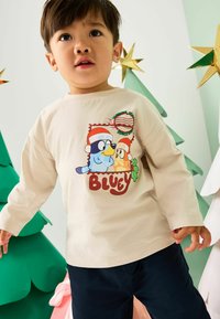 Młody chłopiec w beżowej koszuli z postaciami z kreskówki Bluey w czapkach Mikołaja, stojący obok zielono-białych dekoracji choinkowych.