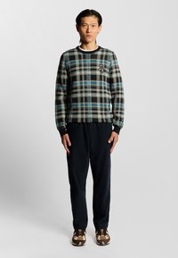 Schwarzer und grauer karierten Pullover mit blauen Akzenten, gerippten Bündchen und Kragen. Kombiniert mit dunklen Hosen und gemusterten braunen Schuhen.