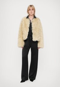 Cremefarbene Jacke aus Kunstfell mit kastiger Form und weiten Ärmeln, getragen über einem schwarzen Top und lockeren schwarzen Hosen. Einfache Knopfdetails sind sichtbar.