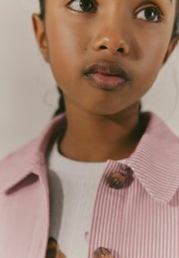 Veste en velours côtelé rose avec un col et des boutons en bois, superposée sur un haut blanc texturé avec un motif floral.