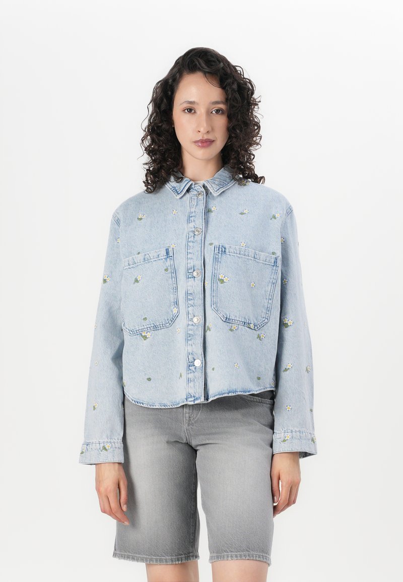 Giacca in denim blu chiaro tagliata sopra il punto vita con due tasche sul petto, bottoni argentati e ricami floreali. Indossata sopra pantaloni corti grigi.