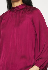 Blusa fucsia con colletto alto, dettagli arricciati sul collo e maniche lunghe e voluminose con polsini elastici. Tessuto morbido.