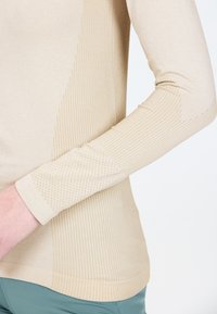 Beige Langarmtop mit glatter Textur, mit gerippten Akzenten an den Seiten und einem gemusterten Bund. Entwickelt für aktive Freizeit.