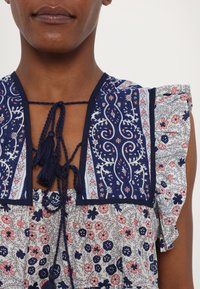 Blusa floral azul marino con mangas acampanadas, que presenta un detalle de lazo con borlas y patrones bordados intrincados en rosa y blanco.