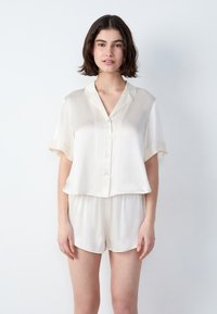 Młoda kobieta z krótkimi ciemnymi włosami, ubrana w luźną, kremową koszulę z krótkim rękawem i pasujące szorty, na jednolitym białym tle.
