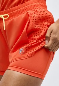 Φωτεινά πορτοκαλί αθλητικά σορτς με διάτρητη δίχτυ πλευρική επιφάνεια και εσωτερική επένδυση συμπίεσης, που κρατιούνται από ένα χέρι κοντά στην τσέπη.