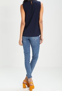 Haut sans manches bleu marine avec un détail de dos en forme de clef, associé à un jean skinny bleu et des sandales à talons carrés jaunes. Tissu lisse, coupe ajustée.