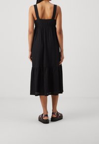 Schwarzes, ärmelloses Kleid mit einem gestuften Rüschensaum, aus strukturiertem Stoff und mit breiten Trägern. Kombiniert mit Riemchensandalen.