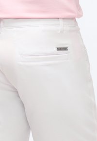 Λευκό παντελόνι με πίσω τσέπη welt και μεταλλική ετικέτα λογότυπου Calvin Klein, φορεμένο με πουκάμισο σε ανοιχτό ροζ χρώμα, μπαλωμένο μέσα.