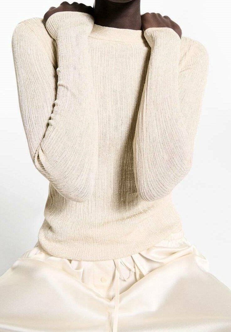Pull beige en maille côtelée avec manches longues, design ajusté et texture subtile ; porté avec un pantalon en satin de couleur claire.