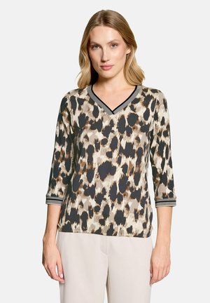 Frau mit blonden Haaren, die ein beige-schwarzes, leopardgemustertes V-Ausschnitt-Oberteil mit Dreiviertelärmeln und hellbeige Hosen trägt.