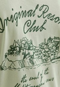 Vaaleansävyinen puuvillapaita, jossa on tummanvihreä grafiikkateksti "Original Resort Club" ja rannikkokylän kuvaus, jossa on rakennuksia ja puita.