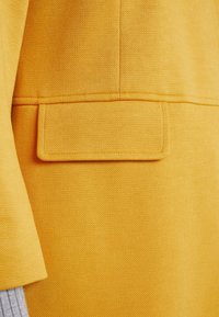 Keltainen teksturoitu kangas, jossa on taskupesä, jossa on siisti ommel. Materiaalissa on kevyt kiilto ja se on rakenteellinen.