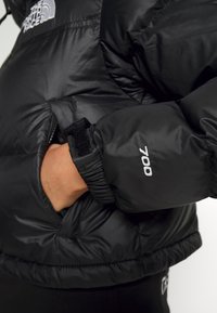 Jachetă puffer neagră cu un finisaj lucios, având manșete elastice și un buzunar. "700" este brodat pe mânecă.