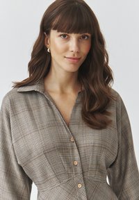 Camicia a quadri marrone e grigia con colletto, chiusura a bottoni e maniche lunghe; il tessuto ha una leggera lucentezza e texture.