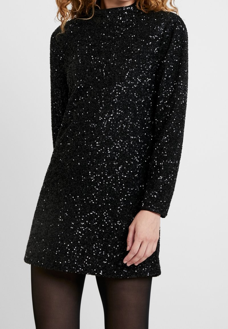Svart paljettklänning med långa ärmar och hög hals. Tyget är texturerat och glänsande, med scatterade, reflekterande paljetter överallt.