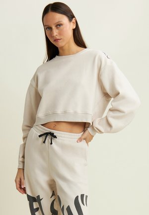 Felpa cropped beige realizzata in tessuto morbido, con vestibilità comoda e maniche lunghe. Coulisse nera sui pantaloni beige abbinati che presentano un testo bold in nero.