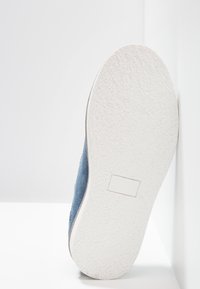 Scarpa in camoscio blu con una tomaia testurizzata e una suola piatta in gomma bianca, caratterizzata da una etichetta rettangolare con il marchio sulla superficie della suola.