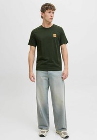 Dunkelgrünes Baumwoll-T-Shirt mit kurzen Ärmeln und einem quadratischen Logopatch auf der Brust, kombiniert mit hellblauen weit geschnittenen Jeans und Sportschuhen.