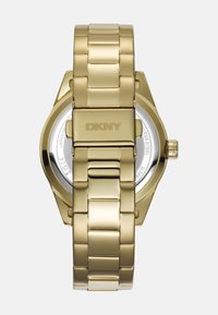 Gouden roestvrijstalen horloge met een gelinkte band, een octagonaal kast en een ronde glasdisplay aan de achterkant met het DKNY-logo.