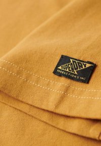 Tela de camiseta amarilla mostaza con una textura suave; presenta una etiqueta negra con el texto amarillo "SUPERDRY" y el logotipo "PRODUCTIONS INC".