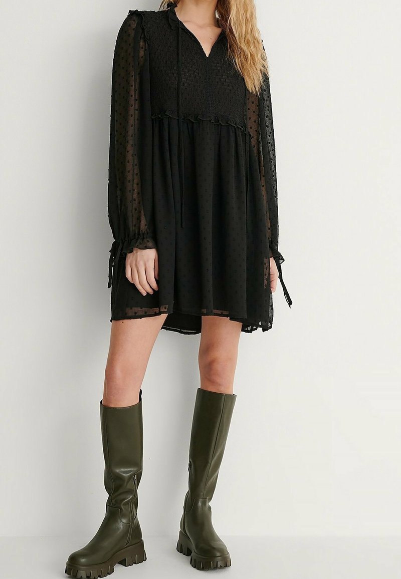 Robe noire à pois avec des manches longues, un corsage smocké, des accents en volants et une jupe évasée. Associée à des bottes vert olive montantes.