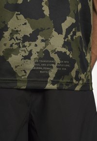 Acercamiento a camisa de camuflaje verde y negro con texto impreso pequeño encima de pantalones negros, brazo parcialmente visible en el lado derecho.