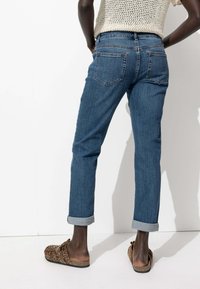 Jean en denim de couleur bleu moyen, présentant une jambe fuselée, un ourlet retroussé et deux poches arrière. Le tissu a une texture lisse avec des coutures subtiles.