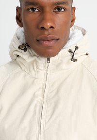 Jachetă din corduroy crem cu guler înalt, finisaj texturat și accesorii din sfoară maro. Captusită cu material fleece de culoare deschisă pentru izolație.