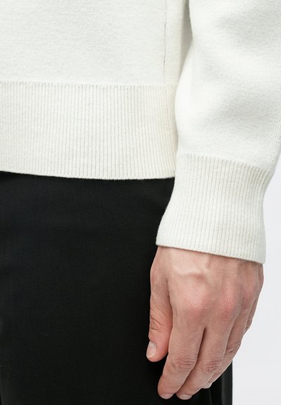 Πουλόβερ από ελαφρύ πλεκτό ύφασμα σε ανοιχτό χρώμα με ριμπ τελείωματα στη μέση και στα μανίκια, συνδυασμένο με παντελόνι από σκούρο ύφασμα. Έμφαση στην υφή και στις αντιθετικές αποχρώσεις.