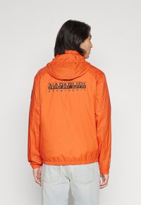 Giacca leggera arancione brillante con cappuccio. Presenta un logo stampato nero sulla parte superiore della schiena e polsini elastici per un design aderente.