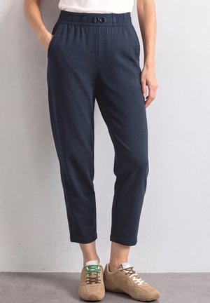 Kvinde iført marineblå cropped bukser med elastisk talje og en dekorativ spænde, kombineret med beige sneakers med ujævne grønne og brune snørebånd.