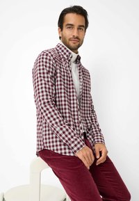 Camicia a quadri rossi e bianchi con maniche lunghe, abbinata a pantaloni in velluto a coste color bordeaux, seduta su una sedia bianca. Sfondo semplice.