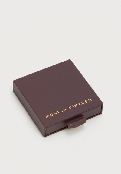 Petite boîte fermée de couleur brun foncé avec languette de tirage et texte doré "Monica Vinader" sur le couvercle, posée sur une surface claire.