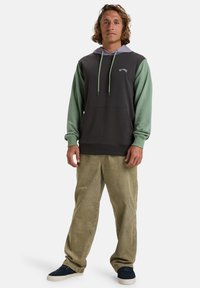 Sudadera con capucha de color gris oscuro en el cuerpo, mangas verdes y acentos morados. Cuenta con un bolsillo canguro y cordones ajustables. Se lleva con pantalones de cordón beige.