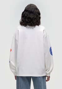 Felpa oversize bianca con spalle abbassate, caratterizzata da una grafica blu su una manica e dettagli rossi sull'altra. Testo sul retro.