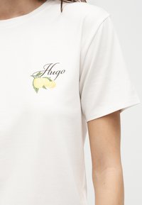 Λευκό πουκάμισο με κοντό μανίκι με τυπωμένη εικόνα δύο κίτρινων λεμονιών και τη λέξη «Hugo» με καλλιγραφικά γράμματα στο αριστερό στήθος.