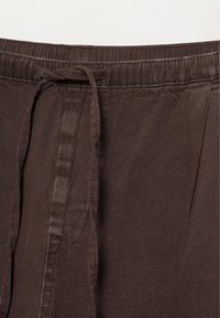 Hnedé látkové nohavice s opaskom na stiahnutie viazaným na uzol a viditeľnými švami vpredu.