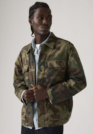 Man met gevlochten haar, gekleed in een camouflagejasje over een licht denim overhemd, kijkt opzij tegen een effen achtergrond.