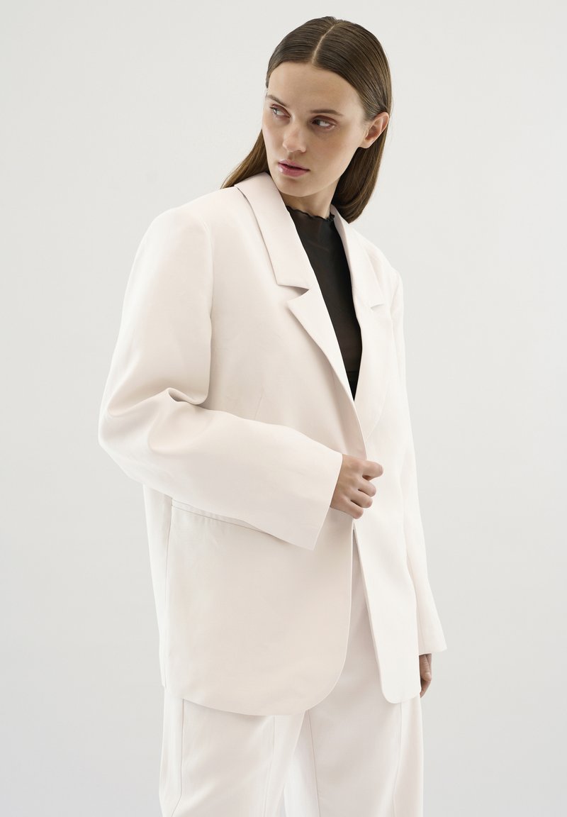 Blazer oversize rosa chiaro con ampi revers, vestibilità morbida e cuciture semplici. Indossato sopra un top nero, su uno sfondo neutro.