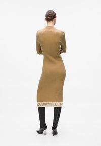 Abito lungo marrone a coste con scollatura a girocollo, silhouette aderente e logo bianco "KARL LAGERFELD" all'orlo; abbinato a stivali neri fino al ginocchio.