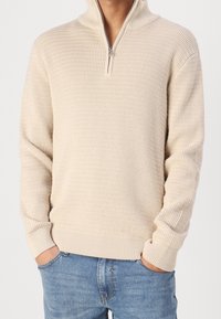 Pull beige à col zippé avec un motif côtelé texturé. Manches longues et taille ajustée, associé à un jean bleu clair.