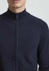 Námornícky sveter na zips s vysokým golierom, ktorý má štruktúrovaný rebrovaný vzor a hladký zips. Materiál sa zdá byť mäkký a priliehavý.